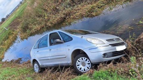 A large silver car mostly on one side next to a water-filled ditch. 