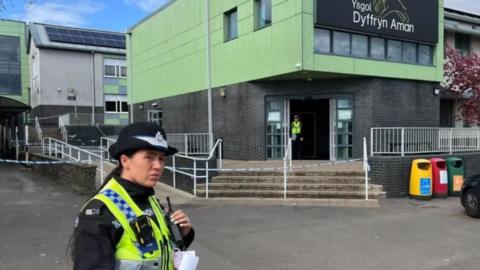 A police officer outside Ysgol Dyffryn Aman in Ammanford, with another office visible in the distance just inside the school and behind police tape. The name of the school is on the building's exterior