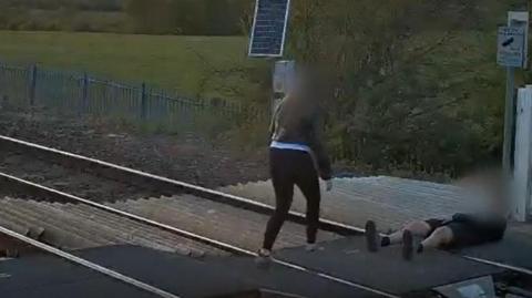 A person in shorts with their face blurred is seen lying on a railway track. A person in black trousers and a jacket with their face blurred is walking over the track towards them.