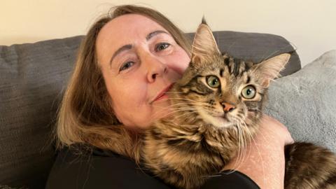 A woman sitting on a grey sofa. She has shoulder-length brown hair and is smiling at the camera. She is holding a brown cat on her shoulder that covers part of her face. The cat has grey eyes and is looking to the right.
