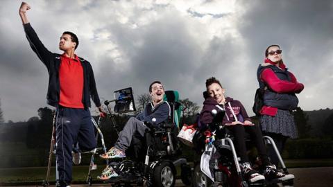 Four people outside, with one person on the left standing using sticks and raising a fist. Two others are seated in wheelchairs, one of which has a communication device attached, and another is stood with their arms crossed and wearing sunglasses. A cloudy sky and trees are visible in the background.
