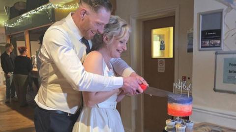 A man and a woman cutting a wedding cake on their wedding day. In the background two people are standing at a bar, with fairy lights hanging from the ceiling.