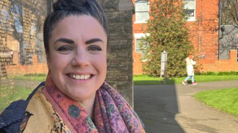 A head and shoulders photo of Katie-Lou Green smiling at the camera. She is wearing a black coat and a multi-coloured scarf. She has black hair which is tied up. She is stood outside in front of a stone building and memorial plaque. 
