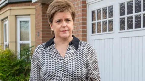 Nicola Sturgeon standing outside her house. She is wearing a black short with white spots
