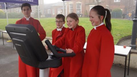 A group of four members of the school choir, dressed in bright red robes with white collars. One of the girls is sitting on the static bike and looking at the electronic screen, while the others stand beside her smiling at encouraging her. The weather is grey, wet and misty. The bike is positioned underneath a blue cabana which says The King's School Gloucester on it.