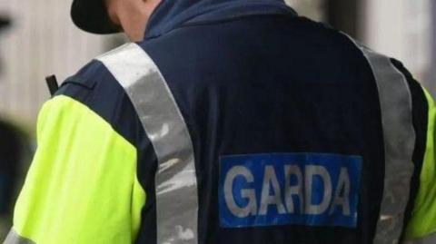 An Garda officer ( Irish police) in uniform is seen from behind. The word garda is spelled out on the back of a high viz jacket, which is partly yellow and has silver reflective bands. Only part of the officers face can be seen under his police cap.