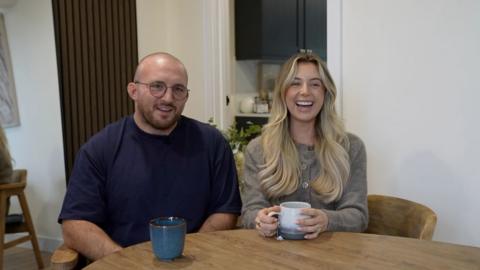 A man and woman stand close together both smiling into camera. The man has a shaved head, is wearing glasses and a dark blue t-shirt. The woman has long blonde hair, blue eyes and is wearing a grey cardigan.