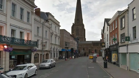 The image shows Broad Street in Hereford. Some cars are parked on the left outsides shops/businesses and there are some empty shops on the right hand side. The street is wide and most building are white/cream in colour. A church tower/spire can be seen at the end of the street.


