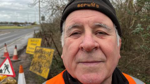 Mark Fishpool, 68, has grey hair hair around his ears, he is wearing a black beany hat with a high-viz orange jacket. Behind him are a selection of old road signs, stands for them, bollards, sandbags and safety lamps.