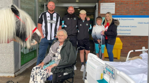 Allison Charlton, an elderly woman, sits in a wheelchair and looks on as a horse called Charlie stands to her side. A hospital bed is to her right and it is outside of a building entrance. Two men and two boys and one girl stand behind the woman.
