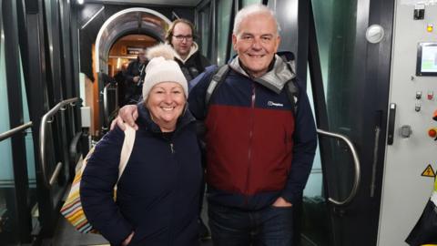 Two passengers disembark from the ferry. The woman on the left is wearing a cream bobble hat and a blue jackert and is carrying a striped bag. The man on her right, who has his arm round her shoulder, is wearing a red and blue jacket. 