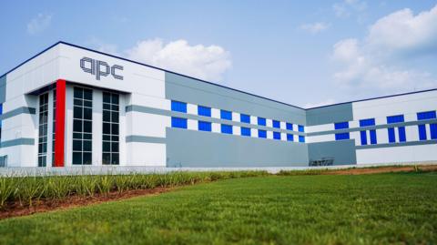 A large factory with APC logo. It is a grey and white building with blue windows. There is a green lawn in front of building
