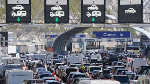 Heavy traffic sits in queues at the Port of Dover check-in area on 22 May 2026, with cars, campervans, vans and lorries filling multiple lanes leading towards border control and ferry processing points. Large overhead electronic signs display lane directions for different vehicle types, while check-in and border control buildings can be seen further ahead beneath the cliffs.