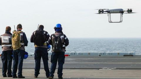 Four men stand in a row wearing black high-viz with 700X written on the back of one of the jackets. They appear to be holding consoles and before them, hovering over the deck of an aircraft carrier with the sea byond, is a Malloy T-150 drone