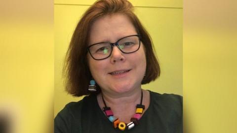 Jo Burridge looking at the camera in front of a green wall wearing a Liquorice Allsorts necklace