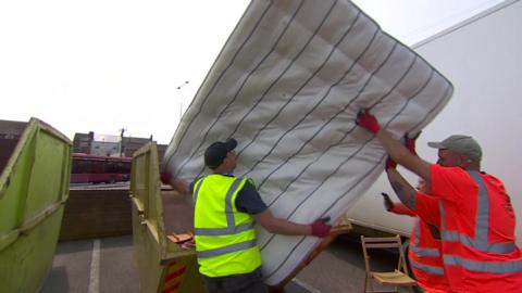 Workers in orange and yellow high-vis vests putting a large mattress into a yellow-coloured skip