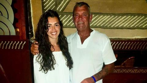 Picture of a white curly long haired brunette woman in a white top, standing next to her father, a white middle aged man with grey hair in a white shirt.