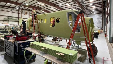 The shell of the body of a plane. It is painted green. There are three red ladders in front of the plane. The two ladders towards the back of the plane have people on them. There are two people inside the plane. On the left there is a person leaning over a work table. The plane is in a airplane hanger. 