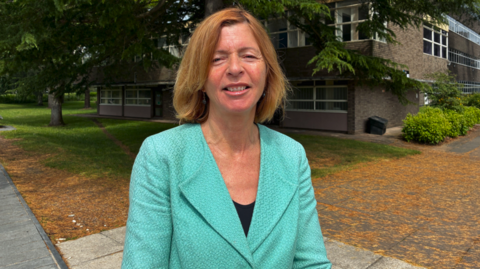 A woman wearing a green jacket stands outdoors in front of trees and a brick building.