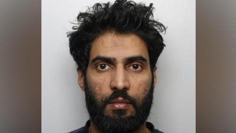 The image is police-style custody or mugshot photograph taken against a plain light-grey background. The picture shows a person facing directly towards the camera, with dark, curly hair and a full beard and moustache.