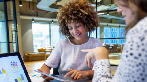 Two women sit opposite each other looking at a laptop screen and an iPad device. They are in an open-plan office with windows behind them as well as chairs and a desk.