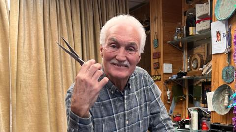 Paul, who has short white hair and a moustache, poses in his salon while holding a pair of scissors. Shelves packed with items can be seen in the background and decorations hanging from the wall.