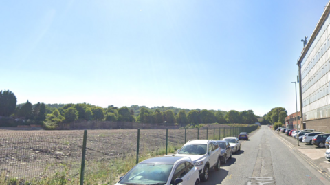 There is brown gravel on the left side of the road which is fenced off with cars parked along the fence. A long, glass and brick building stands on the right hand side with cars parked outside.