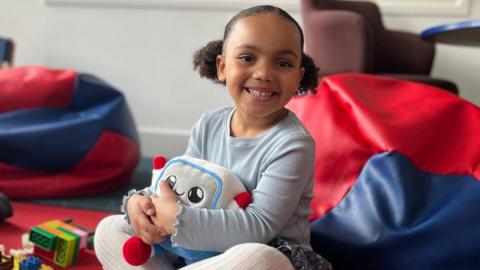Picture of a young girl hugging Gabbo an AI-powered toy. She is sat, smiling among soft seating and other toys.