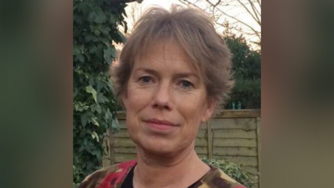A woman with short dark blonde hair is pictured in a garden setting with a wooden fence behind her and a large plant over her right shoulder. 