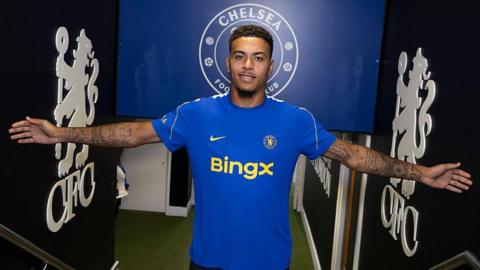 Morgan Rogers, wearing a Chelsea kit, stands with arms outstretched inside the entrance to the pitch at Stamford Bridge