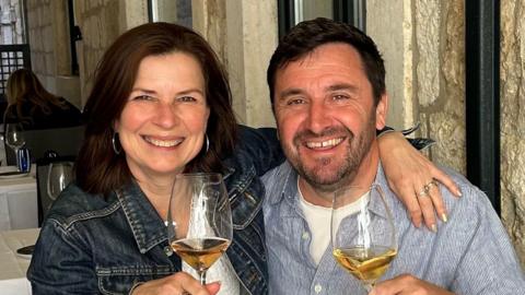 A woman and a man sit side-by-side in a restaurant. The pair are holding a wine glass each containing wine. They are smiling a tthe camera. The woman has placed her arm around the man.