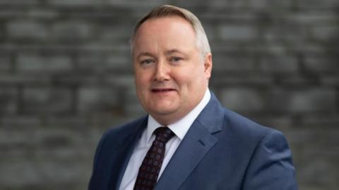 Darren Millar, wearing a blue suit, burgundy and blue polka dot tie and white shirt, in front of the Senedd's slate facade.