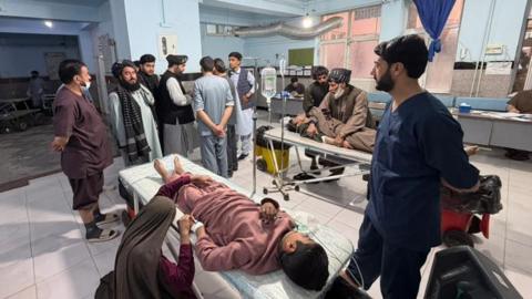 A young male is seen lying down on a stretcher in a hospital ward, hooked up to a drip, as a woman kneels by his side and holds his hand. A man in scrubs is seen stood next to the stretcher looking on at a group on men gathered at the end of the stretcher, while another person can be seen lying on a stretcher in the background
