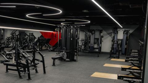 A gym room with squat racks on one wall and bench presses in front of a mirror wall.