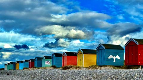 Brightly-coloured beach huts - red, blue, yellow and green - under a cloudy blue sky.