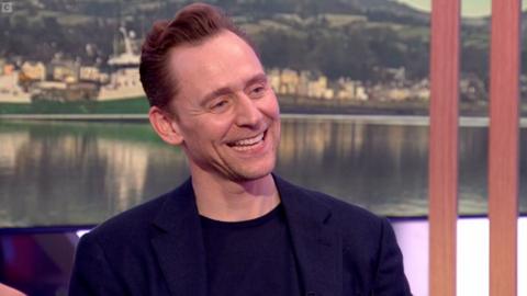 Tom Hiddleston smiles. He has swept back hair and wears a blue blazer and black T-shirt.