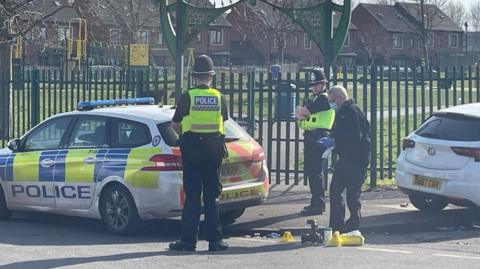 Police officers and two cars at the entrance to a small park with a green metal fence surrounding it and an arched gateway