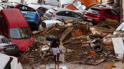 A boy is seen walking among muddy debris and a pile of cars in Valencia 