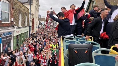 A person holding a silver trophy on an open-top bus. There are hundreds of people in the street.