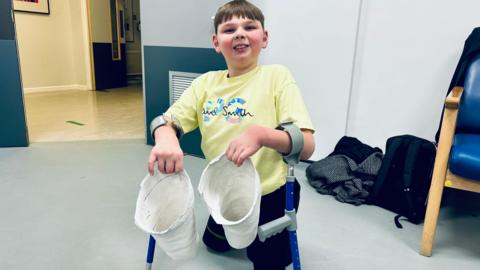 Tony Hudgell pictured at the Bowley Close Rehabilitation Centre in Crystal Palace holding both of his prosthethic limbs. Tony is wearing a yellow t-shirt and black trousers and is using blue crutches with grey arm holds and handles.