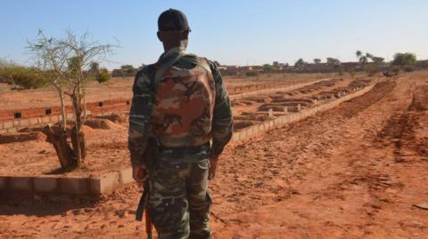 An armed soldier wearing military fatigue looks at the graves in open area 