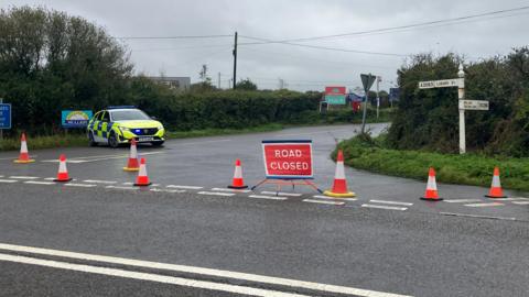 A police car at the junction of a road which is closed with a road closed sign on it following a crash.