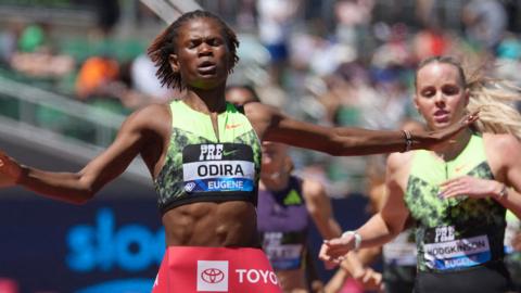 Kenya's Lilian Odira closes her arms and spreads her arms wide to either side as she crosses the line to defeat Britain's Keely Hodgkinson to win the women's 800m at the Eugene Diamond League. The two athletes are seen from waist up, with Odira wearing red shorts and a green running vest which is cut off to reveal her stomach while Hodgkinson wears a similar green zip-up once-piece vest which fades to black around the waist. Some other runners and the crowd are blurred in the background