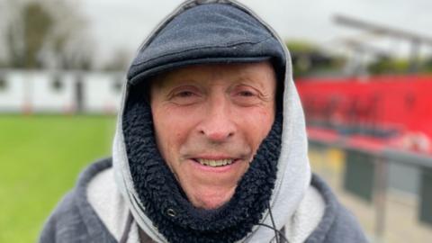 Steve Day standing on a football pitch wearing a hoodie and a cap. He is wearing a jumper underneath his hoodie. He is wearing a cap and has the hood of his hoodie over his head.