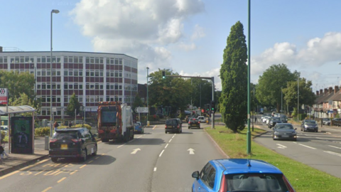 A multi-lane road, with vehicles travelling in both directions. There is a large office block to the top left of the image, and traffic lights straight ahead.