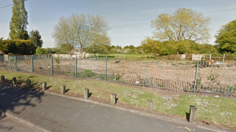 An area behind green fencing, scraped back to the earth, with trees in the distance