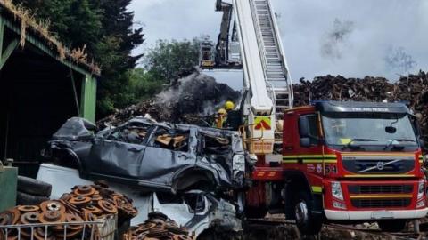 Fire service vehicle backed up to vast piles of scrap metal with a grey crushed car to the left