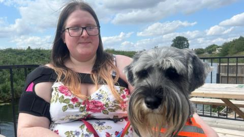 Elanor Maxey and her guidance dog Genie. Eleanor is to the left of the frame and she is wearing a white floral dress, embossed with pink roses. On her right upper arm she is wearing a black and pink support bandage which covers the part of her arm she said was injured in the attack. She has brown straight hair which is lighter at the tips and is wearing black glasses. Her dog, Genie, is sat beside her, she has fluffy grey hair which has been recently cut and a distinct beard which flows from the top of her nose. Her nose is black and shiny. Elanor and Genie are sat on a wooden bench in front of a river. 