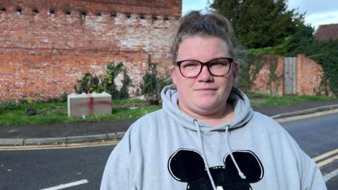 A woman with her hair in a messy bun, glasses and a grey hoodie with a Mickey Mouse outline on it stands on a pavement. On the other side of the road, a concrete block with a red cross spray painted on it has been placed on a grass verge. There is a red brick wall with ivy growing all over it.