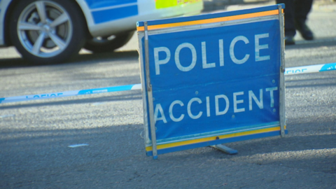 A blue sign on the road reads "Police Accident". The lower part of a police car and police tape can be seen in the background. 